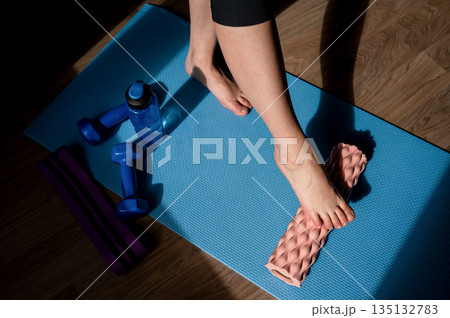 Female legs on foam roller doing myofascial massage with dumbbells and water bottle nearby Female legs on foam roller doing myofascial massage with dumbbells and water bottle nearby 135132783