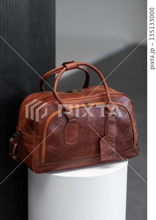 Luxury cognac leather weekender travel bag on a white cylindrical pedestal in a minimalist modern studio with architectural textures. 135133000