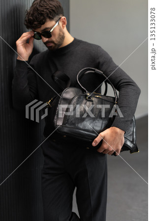Confident man in sunglasses and charcoal knitwear holding a black leather briefcase in a modern minimalist interior. Confident man in sunglasses and charcoal knitwear holding a black leather briefcase in a modern minimalist interior. 135133078