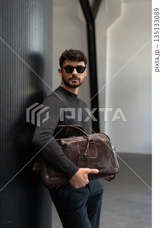 Confident man in sunglasses and charcoal knitwear holding a dark brown leather briefcase while leaning against a textured pillar in a modern minimalist interior. Confident man in sunglasses and charcoal knitwear holding a dark brown leather briefcase while leaning against a textured pillar in a modern minimalist interior. 135133089