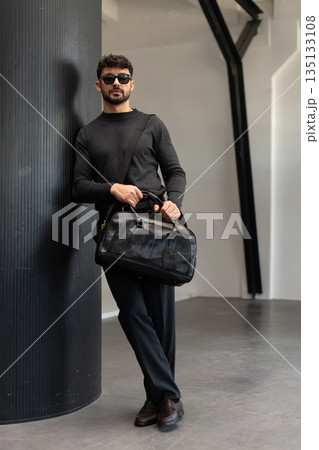 Confident man in sunglasses and charcoal knitwear holding a black leather briefcase while leaning against a textured pillar in a modern minimalist interior. 135133108