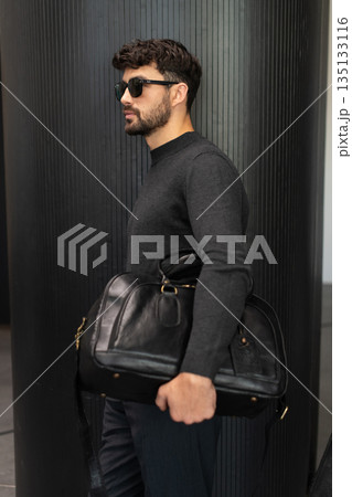 Confident man in sunglasses and charcoal knitwear holding a black leather briefcase while leaning against a textured pillar in a modern minimalist interior. Confident man in sunglasses and charcoal knitwear holding a black leather briefcase while leaning against a textured pillar in a modern minimalist interior. 135133116
