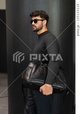 Confident man in sunglasses and charcoal knitwear holding a black leather briefcase while leaning against a textured pillar in a modern minimalist interior. Confident man in sunglasses and charcoal knitwear holding a black leather briefcase while leaning against a textured pillar in a modern minimalist interior. 135133119