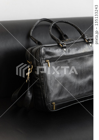 A professional black leather briefcase with dual handles and adjustable strap sits on a sleek black leather sofa in a minimalist studio setting. 135133243