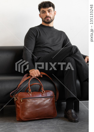 A professional male model in charcoal knitwear and tailored trousers sits on a black leather sofa holding a premium light brown leather briefcase. A professional male model in charcoal knitwear and tailored trousers sits on a black leather sofa holding a premium light brown leather briefcase. 135133248