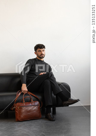 A professional male model in charcoal knitwear and tailored trousers sits on a black leather sofa holding a premium light brown leather briefcase. A professional male model in charcoal knitwear and tailored trousers sits on a black leather sofa holding a premium light brown leather briefcase. 135133251