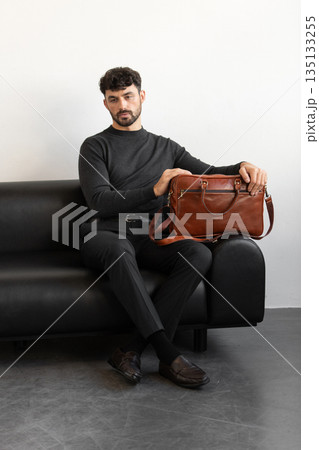 A professional male model in charcoal knitwear and tailored trousers sits on a black leather sofa holding a premium light brown leather briefcase. 135133255