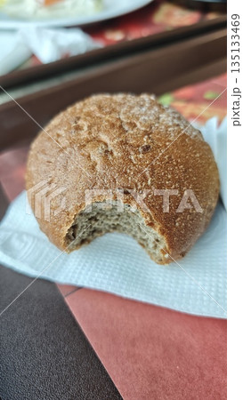 Whole grain bread roll with a bite taken out placed on napkin on cafeteria table. Simple food moment concept representing everyday meal break and casual eating lifestyle 135133469
