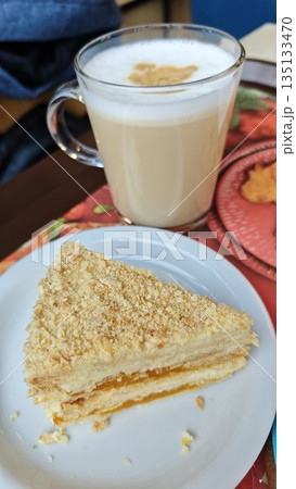 Layered cream cake slice with crumb topping served on white plate alongside glass of latte. Cozy dessert break concept with comfort food enjoyment and relaxed cafe lifestyle 135133470