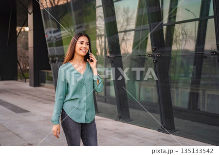 Smiling businesswoman walking near office building and talking on smartphone Smiling businesswoman walking near office building and talking on smartphone 135133542