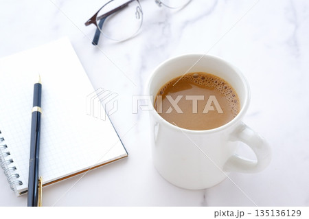 コーヒーを飲みながら、リング式のメモ帳とペンでデスクワークをしているイメージ 135136129