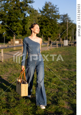 Young adult beautiful woman wear stylish jeans one-shoulder denim jumpsuit have fun walks outdoors warm sunny day holding straw bag hat hand. Female person carefree smiles enjoying countryside nature 135136503