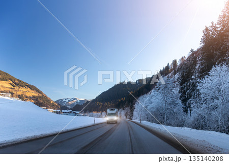 Winter road scene with truck driving through alpine valley surrounded by snowy fields and frosted forest under clear blue sky. Mountain landscape, cold weather, transport route, calm rural atmosphere 135140208