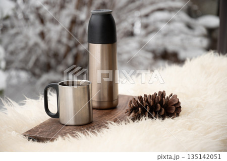 Stainless steel thermos and metal mug on wooden board with pine cone outdoors. Winter picnic and hot drink concept in cold weather. Stainless steel thermos and metal mug on wooden board with pine cone outdoors. Winter picnic and hot drink concept in cold weather. 135142051