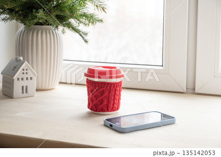 Reusable coffee cup in red knitted sleeve and smartphone on windowsill. Cozy winter morning and remote work lifestyle concept. Cozy home interior. Reusable coffee cup in red knitted sleeve and smartphone on windowsill. Cozy winter morning and remote work lifestyle concept. Cozy home interior. 135142053