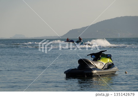 Two jet skis carrying tourists glide across the sea. One jet ski awaits tourists. 135142679