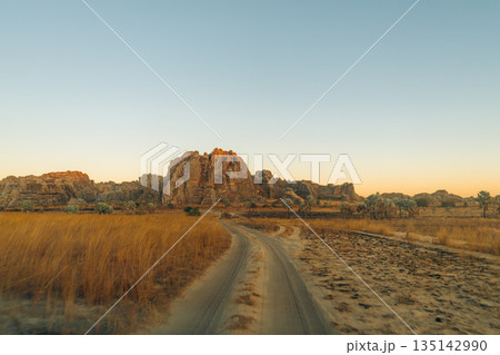 Isalo, Madagascar: Dirt road winding through savanna towards sunlit rock formations 135142990