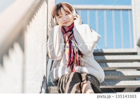 階段に座ってヘッドホンで音楽を聴く若い女性 階段に座ってヘッドホンで音楽を聴く若い女性 135143303