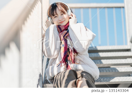 階段に座ってヘッドホンで音楽を聴く若い女性 135143304