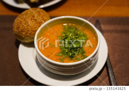 A Delicious Bowl of Lentil Soup Topped with Fresh Herbs Accompanied by a Soft Bread Roll on a Cozy Dining Setting 135143825