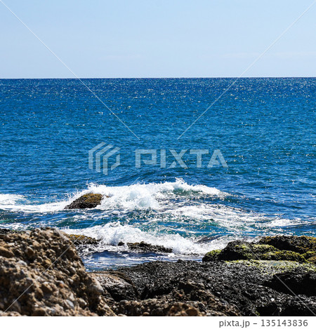 A Tranquil Coastal Scene Featuring Gentle Waves Crashing Against Rugged Rocks Under a Clear Blue Sky, Illustrating Nature's Beauty and Serenity 135143836