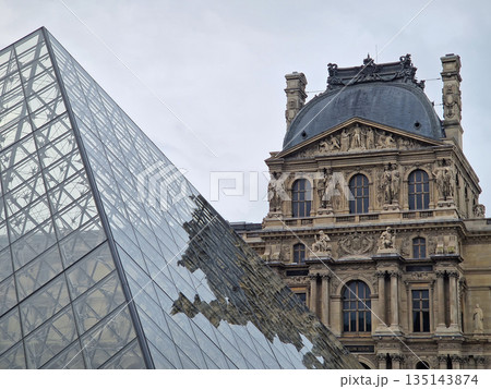 Paris, France - December 10, 2025: The ornate Renaissance facade and the iconic glass Pyramid of the Louvre Museum with multiple tourists scattered across the vast cobblestone courtyard 135143874