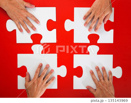 Four hands positioning white puzzle pieces on a red background Four hands positioning white puzzle pieces on a red background 135143969