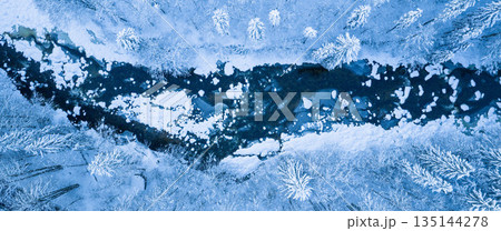 Aerial top down view of a snow covered winter forest with a frozen stream cutting through frosted pine trees. Minimal, serene winter landscape captured by drone in cold blue tones. Aerial top down view of a snow covered winter forest with a frozen stream cutting through frosted pine trees. Minimal, serene winter landscape captured by drone in cold blue tones. 135144278