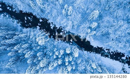Aerial top down view of a snow covered winter forest with a frozen stream cutting through frosted pine trees. Minimal, serene winter landscape captured by drone in cold blue tones. Aerial top down view of a snow covered winter forest with a frozen stream cutting through frosted pine trees. Minimal, serene winter landscape captured by drone in cold blue tones. 135144280