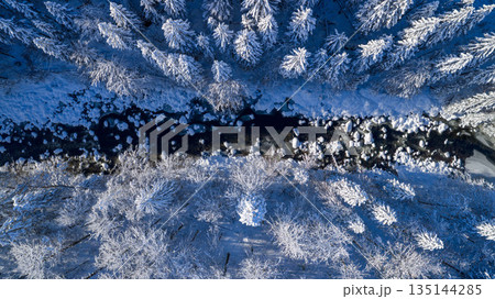 Aerial top down view of a snow covered winter forest with a frozen stream cutting through frosted pine trees. Minimal, serene winter landscape captured by drone in cold blue tones. Aerial top down view of a snow covered winter forest with a frozen stream cutting through frosted pine trees. Minimal, serene winter landscape captured by drone in cold blue tones. 135144285