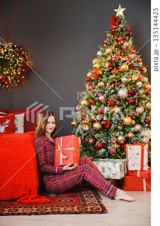 Girl sitting by Christmas tree with gift in living room 135144425