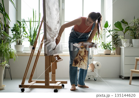 Skillful woman artist enjoying painting picture on easel with paintbrush at modern creative loft. Skillful woman artist enjoying painting picture on easel with paintbrush at modern creative loft. 135144620