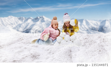 Playful artwork of two girls building snow structure together in mountains. 135144971