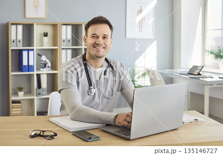 Portrait of a young happy male doctor or medical nurse in uniform sitting at the desk in his office Portrait of a young happy male doctor or medical nurse in uniform sitting at the desk in his office 135146727