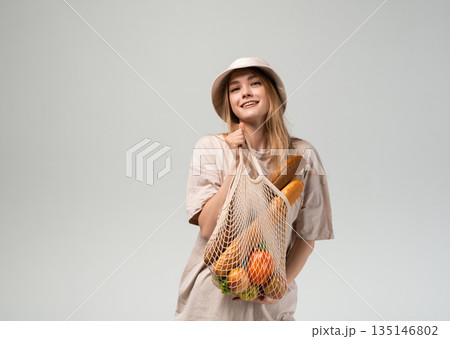 Smiling young woman in bucket hat holding reusable mesh grocery bag with bread and fresh produce, eco lifestyle portrait Smiling young woman in bucket hat holding reusable mesh grocery bag with bread and fresh produce, eco lifestyle portrait 135146802