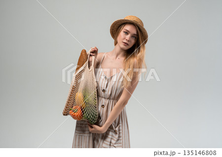 Stylish young woman holding eco friendly grocery bag with fresh produce, straw hat and summer dress isolated on gray background 135146808