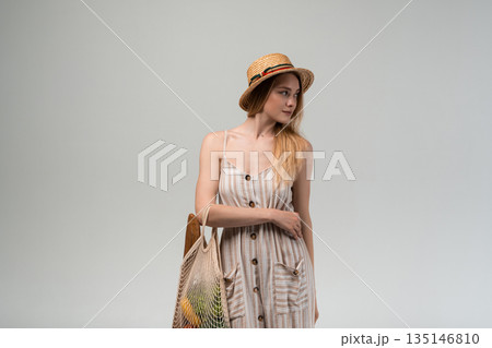 Young woman in straw hat holding eco shopping bag with groceries looking to the side on light background Young woman in straw hat holding eco shopping bag with groceries looking to the side on light background 135146810