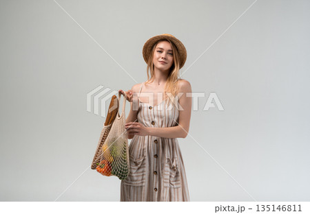 Young woman with eco shopping bag and fresh groceries wearing summer dress and straw hat isolated on light background Young woman with eco shopping bag and fresh groceries wearing summer dress and straw hat isolated on light background 135146811