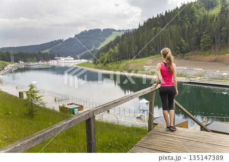 Back view of a young sportive woman in fitness clothes standing outdoors near big lake in mountains enjoying nature view. Back view of a young sportive woman in fitness clothes standing outdoors near big lake in mountains enjoying nature view. 135147389