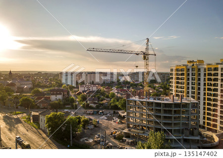 Apartment or office tall building under construction. Brick walls, glass windows, scaffolding and concrete support pillars. Tower crane on bright blue sky copy space background. Apartment or office tall building under construction. Brick walls, glass windows, scaffolding and concrete support pillars. Tower crane on bright blue sky copy space background. 135147402