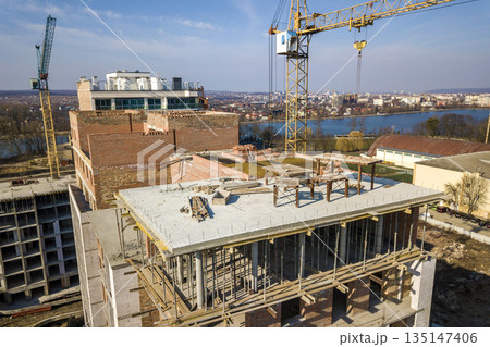 Apartment or office tall building under construction, top view. Brick walls, scaffolding and concrete support pillars. Tower crane on bright blue sky copy space background. Drone aerial photography. Apartment or office tall building under construction, top view. Brick walls, scaffolding and concrete support pillars. Tower crane on bright blue sky copy space background. Drone aerial photography. 135147406