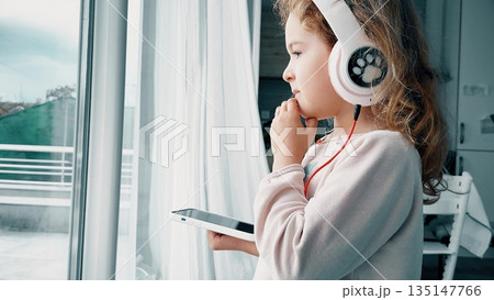 Child listening to music with headphones at home by window 135147766