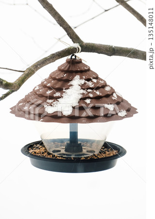 A plastic bird feeder with seeds and bird food hangs from a tree. Caring for birds in winter A plastic bird feeder with seeds and bird food hangs from a tree. Caring for birds in winter 135148011