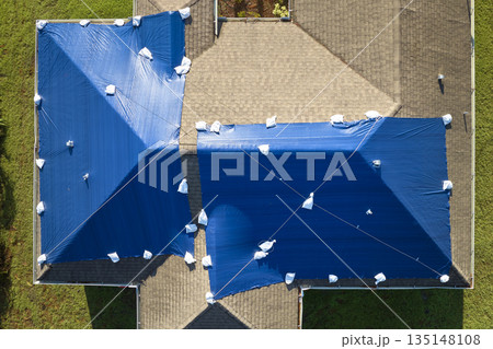 Aerial view of damaged in hurricane Ian house roof covered with blue protective tarp against rain water leaking until replacement of asphalt shingles Aerial view of damaged in hurricane Ian house roof covered with blue protective tarp against rain water leaking until replacement of asphalt shingles 135148108