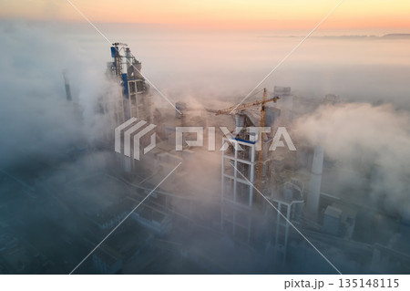 Aerial view of cement factory with high concrete plant structure and tower crane at industrial manufacturing site on foggy evening. Production and global industry concept. 135148115