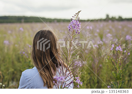 Young beautiful blond woman in purple shirt from behind walking in the meadow among flowers of fireweed 135148363
