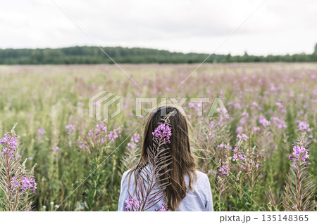 Purple flower of fireweed close-up on the background of blond hair of a young woman in light purple shirt from behind on a meadow 135148365