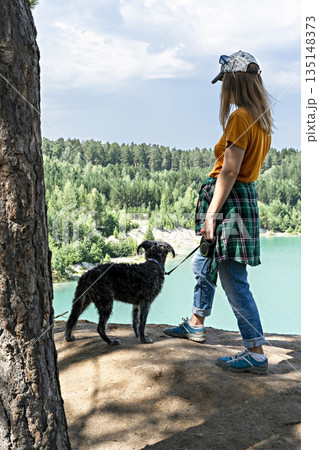 Rear view of young blond woman traveler in a cap and ginger t-shirt standing on the cliff of blue turquoise lake or river and coniferous forest with fluffy dog , travel with pets beautiful landscape 135148373