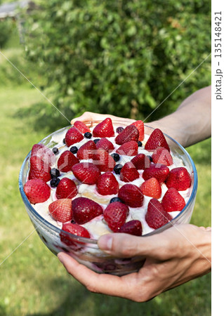 Homemade dessert with strawberries and blueberries and coconut cream in a glass dish in hands on background of green plants outside in summer, vegan cake 135148421