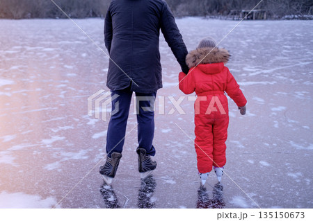 A girl in a red winter jumpsuit learns to skate while holding her dad's hand. Ice skating rink on a frozen lake in a frosty winter A girl in a red winter jumpsuit learns to skate while holding her dad's hand. Ice skating rink on a frozen lake in a frosty winter 135150673